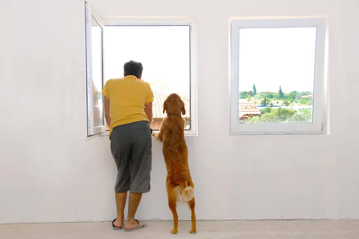 A man standing with his dog looking out his window.